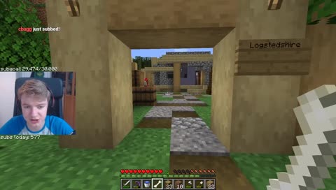 This is a screenshot of Tommy's stream. It shows the inside of Logstedshire. Tommy is stepping through the doorframe into the camp and following a path made out of dirt and gravel. Ghostbur's small house sits in the back of the camp, made out of birch, cobblestone, and blue terracotta. There is a pile of barrels to his left. Tommy holds a bone in his hand. His facecam shows him talking mid-sentence.